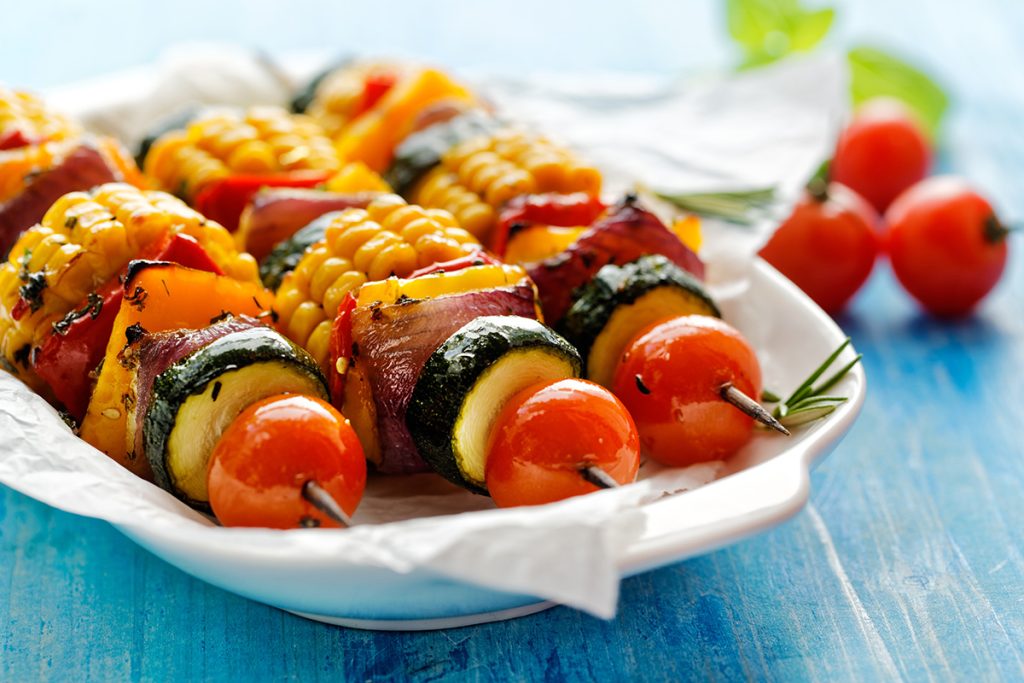 Three vegetarian skewers that consist of cherry tomatoes, zucchinis, red onions, red pepper and corn on the cob that has been sliced up 