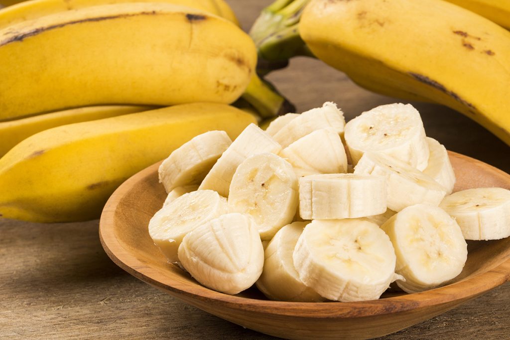 Sliced bananas in a wooden bowl with banana fruits in laying in the background next to the bowl