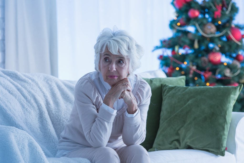 A sad older woman sitting on the couch with a christmas tree in the background