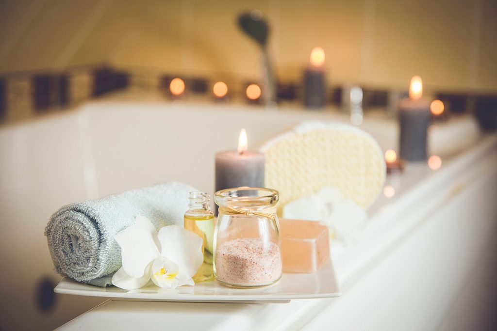 Close up of a tray with towels, soap, bath salts, a flower, and essential oils displayed on the edge of a tub with candles lit all around it