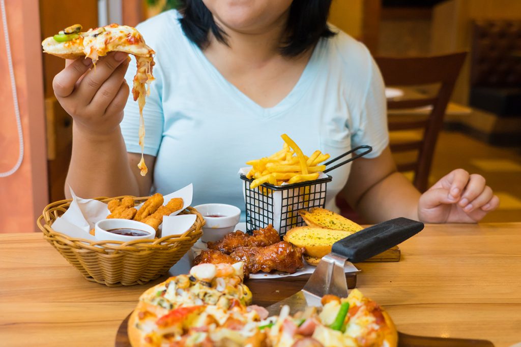 Woman picking up a slice of pizza during a meal that consist of: chicken wings, french fries, chicken tenders and garlic bread displayed in front of her 