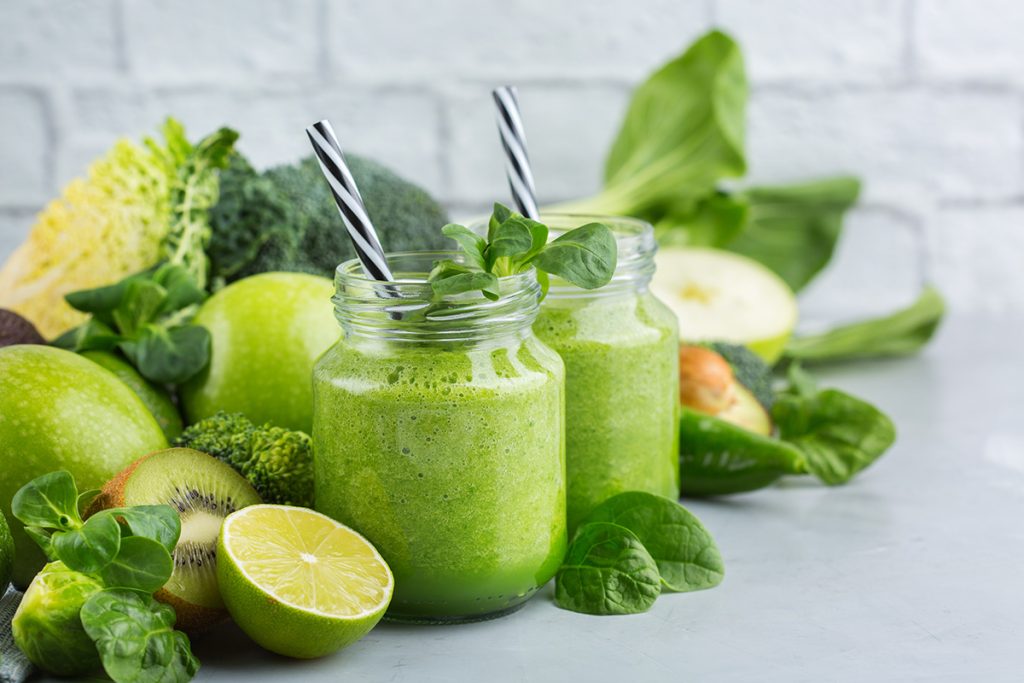Two green smoothies next to a pile of green fruits and vegetables