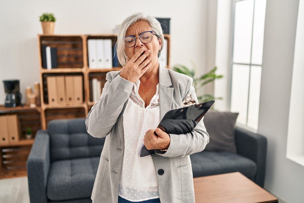 Older woman yawning while holding a clipboard in a living room