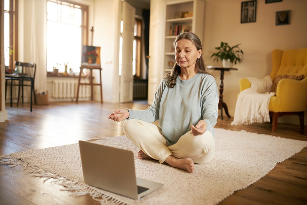 Older woman meditating in living room with laptop