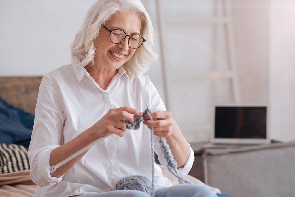 Older woman smiling and knitting in bedroom