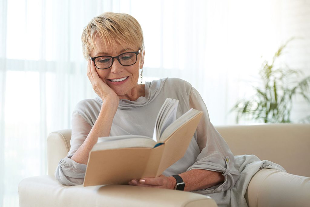 Senior blond woman in glasses resting on sofa and reading a book