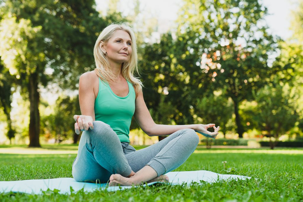 mature woman in sporty outfit relaxing meditating feeling zen-like on fitness mat in public park outdoors. Healthy active lifestyle