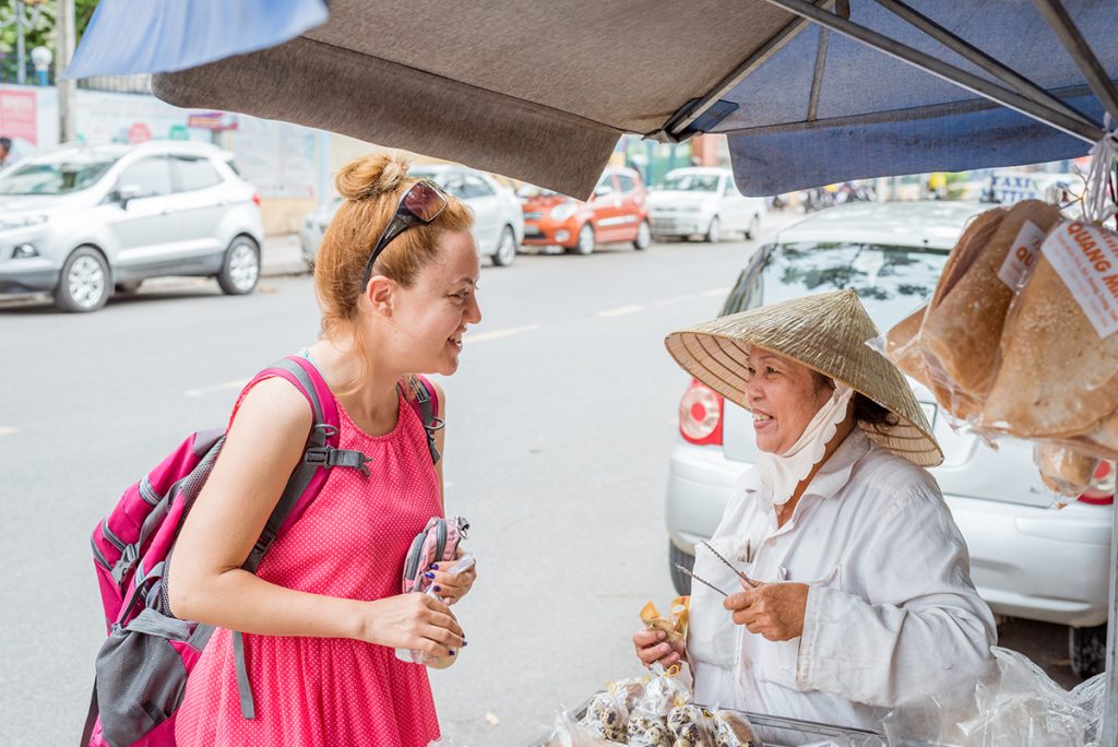 a solo European traveler and a street dessert vendor, a Vietnamese lady in an Asian conical hat, happily look at each other after completing the shopping interaction.
