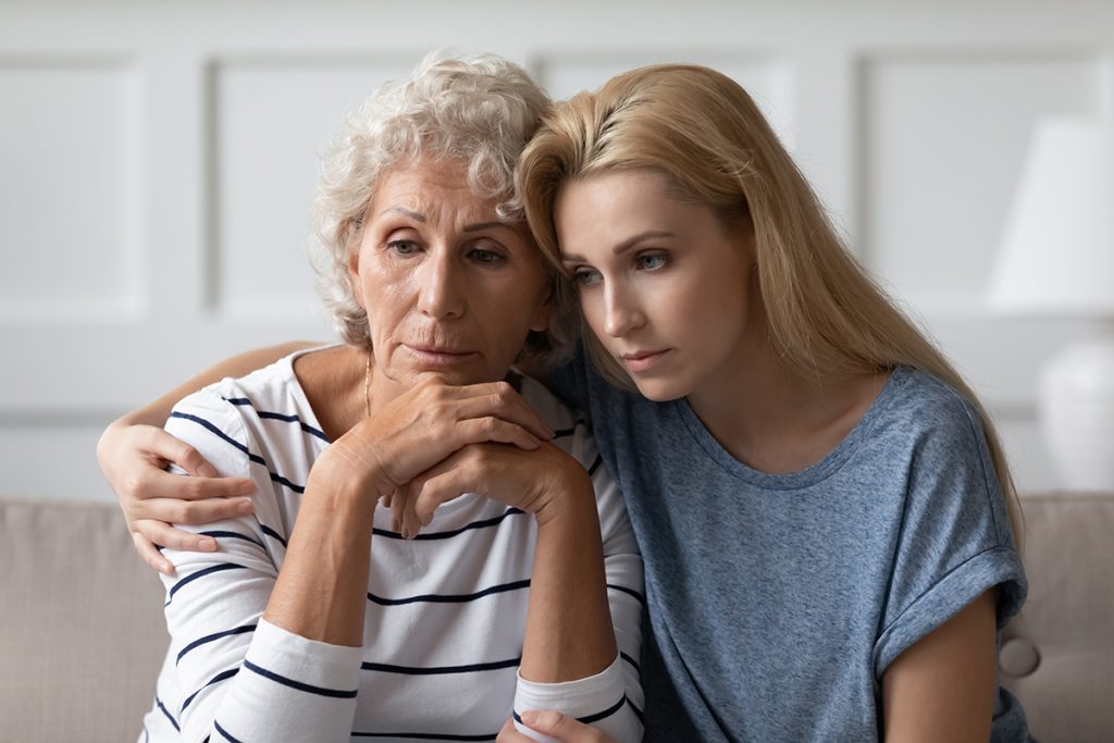 Sad young adult woman grown up daughter or grandkid sitting on sofa hugging desperate, grieving, frustrated elderly mom or grandma having problems with mental health