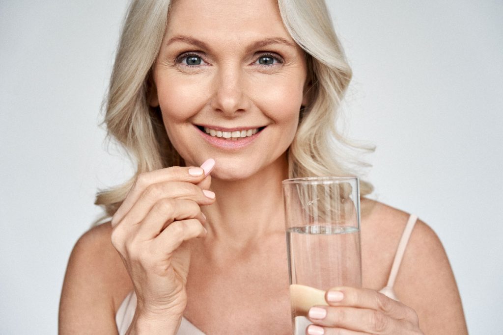 Smiling happy healthy middle aged 50s woman holding glass of water taking dietary supplement vitamin pink pill isolated on white background.