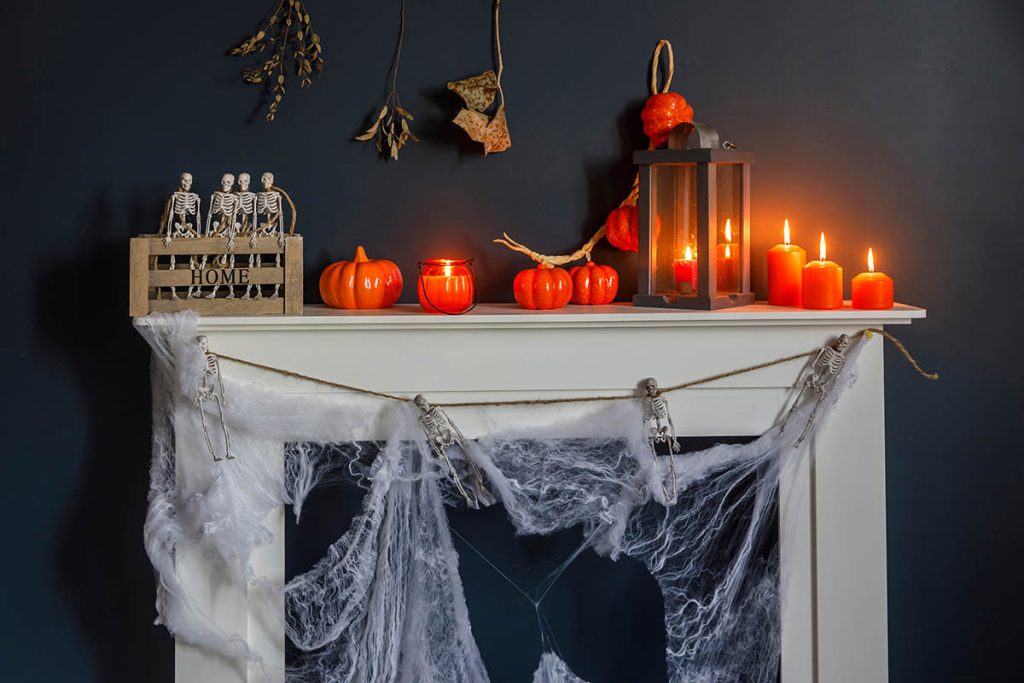 Halloween home decoration. Plastic toy skeletons in a wooden box on the fireplace against a dark blue wall. A garland of skeletons. Cobweb on the dresser. Orange candles and lantern.