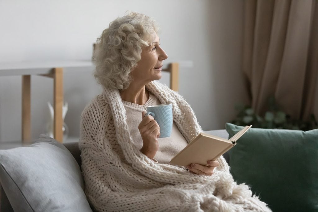middle aged woman wrapped warm blanket sitting on cozy couch, looking to aside, satisfied mature female drinking tea or coffee, reading book, enjoying weekend, relaxing at home