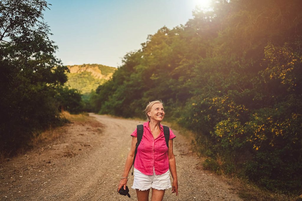 Excited happy senior woman backpacker tourist walking in summer forest road outdoors at sunset time. Old slim lady traveling with photo camera
