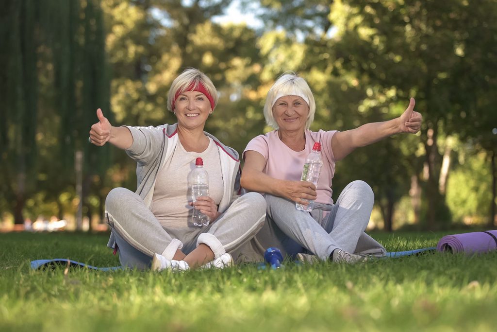 Two women showing thumbs up sitting on grass after doing exercises, positive