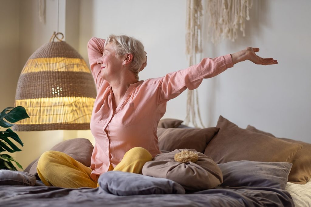 senior woman wearing pyjama smiling in bright living room stretching waking up.