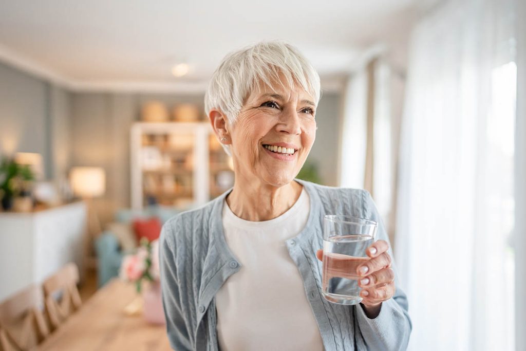Close up portrait of one senior woman with short hair happy smile positive emotion copy space standing at home indoor gray white hair hold glass of water