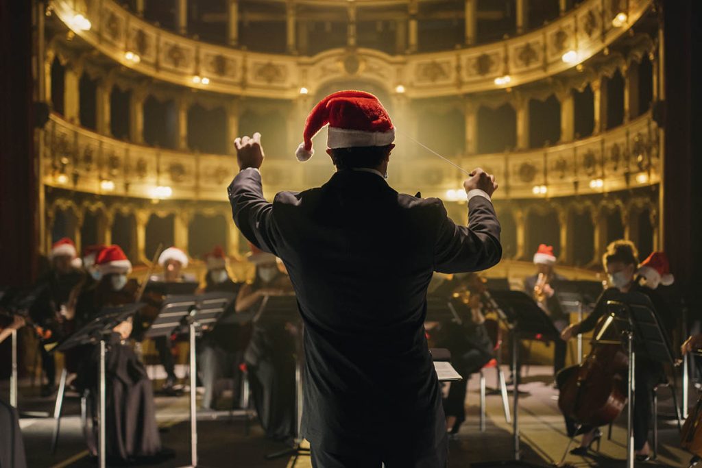 Cinematic shot of Conductor Directing Symphony Orchestra with Performers All Wearing Santa Hats Playing Violins, Cello and Trumpet on Classic Theatre with Curtain Stage During Christmas Music Concert