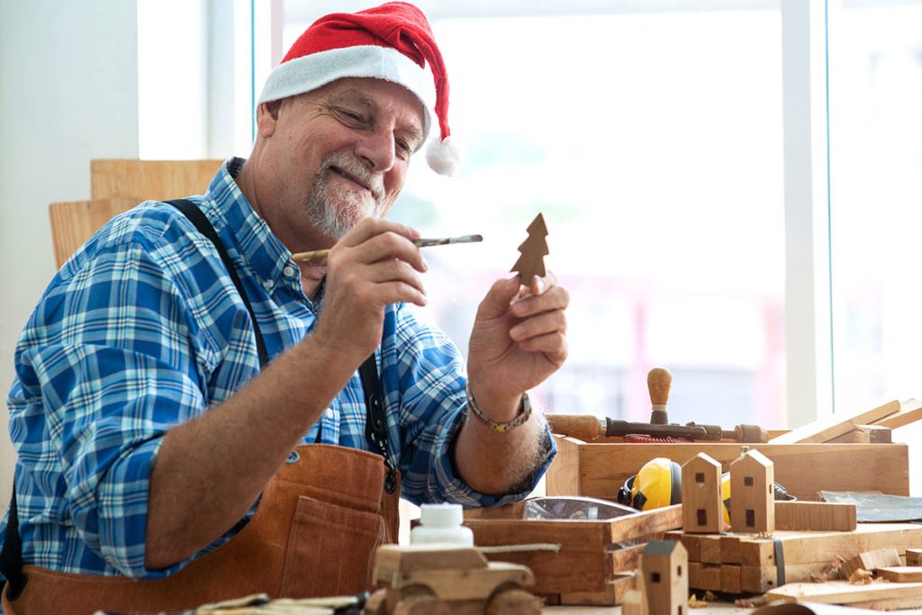 man wearing Santa hat holds and paints small wooden Christmas tree, prepare toys ready for Christmas
