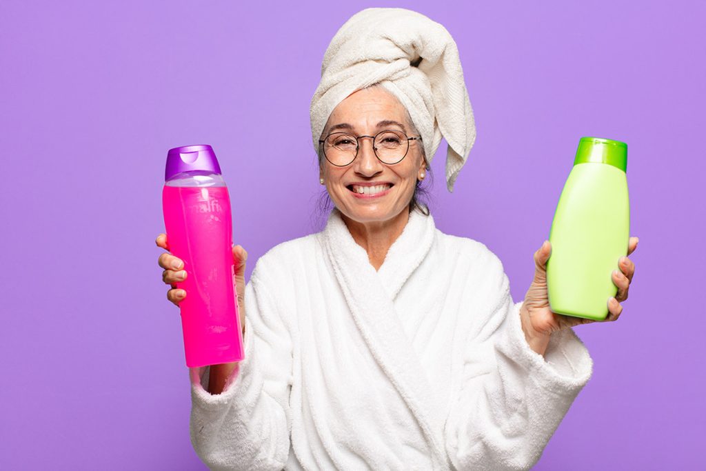 older woman in robe and towel holding shampoo and conditioner smiling