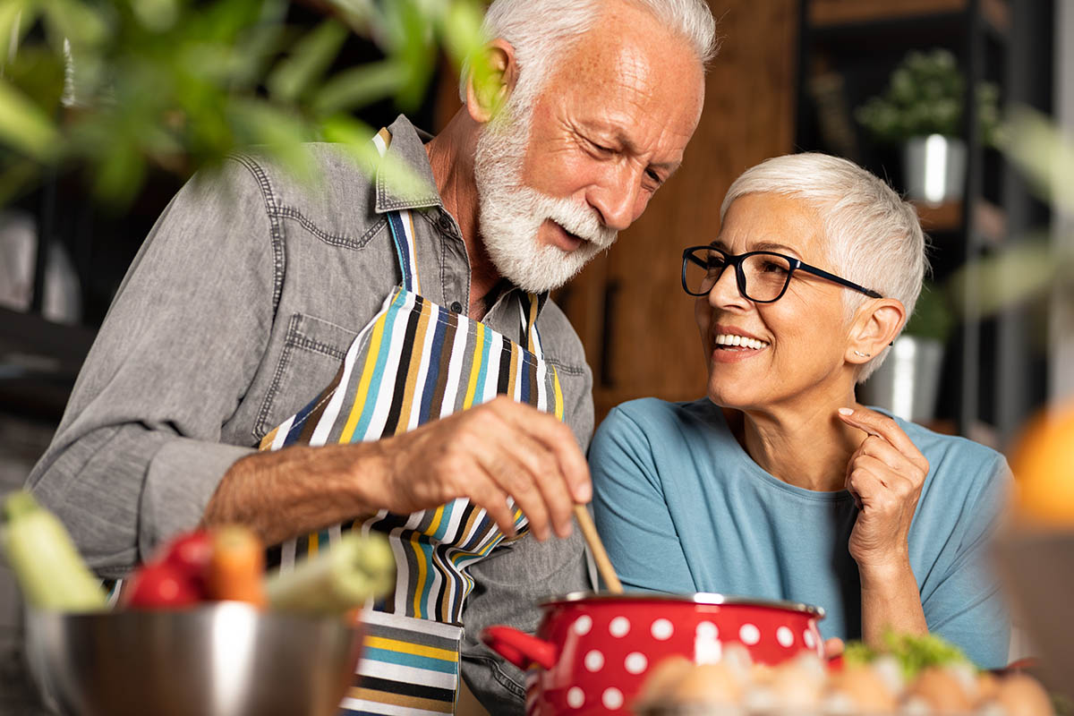 smiling senior couple standing close together, caringly and laughing, cooking at kitchen table