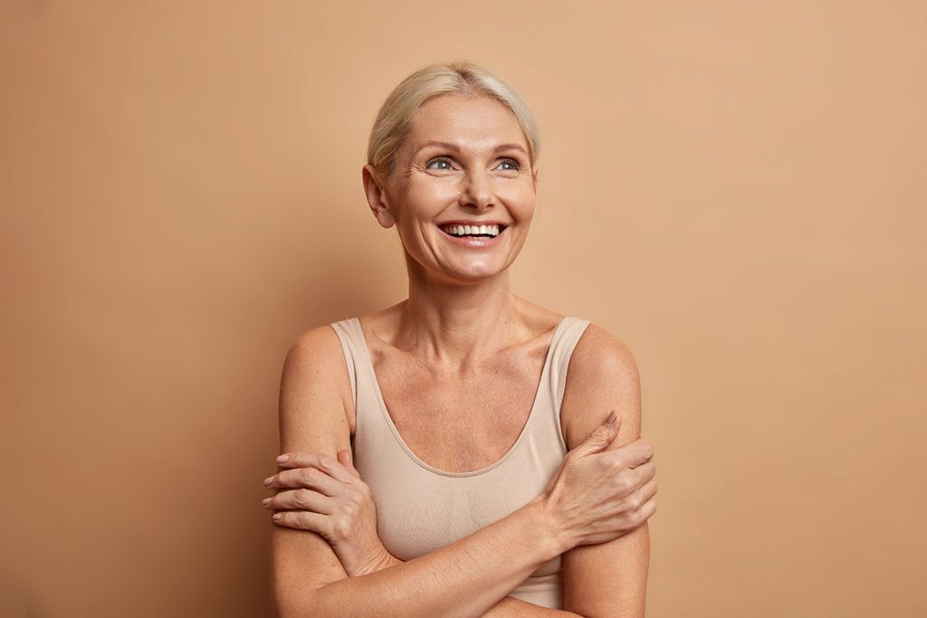 mature woman looks gladfully above keeps arms foded has well cared complexion healthy skin white teeth isolated over brown background. 