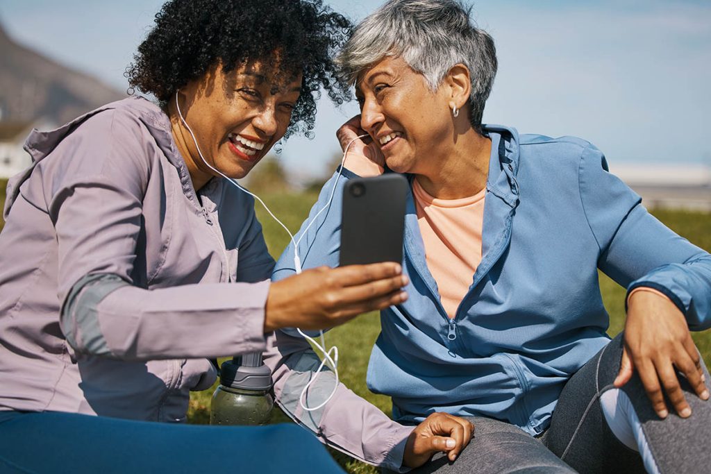 Phone, fitness and music with senior friends on the grass outdoor taking a break from their workout routine. Exercise, smile and elderly people streaming audio while laughing on a field for wellness