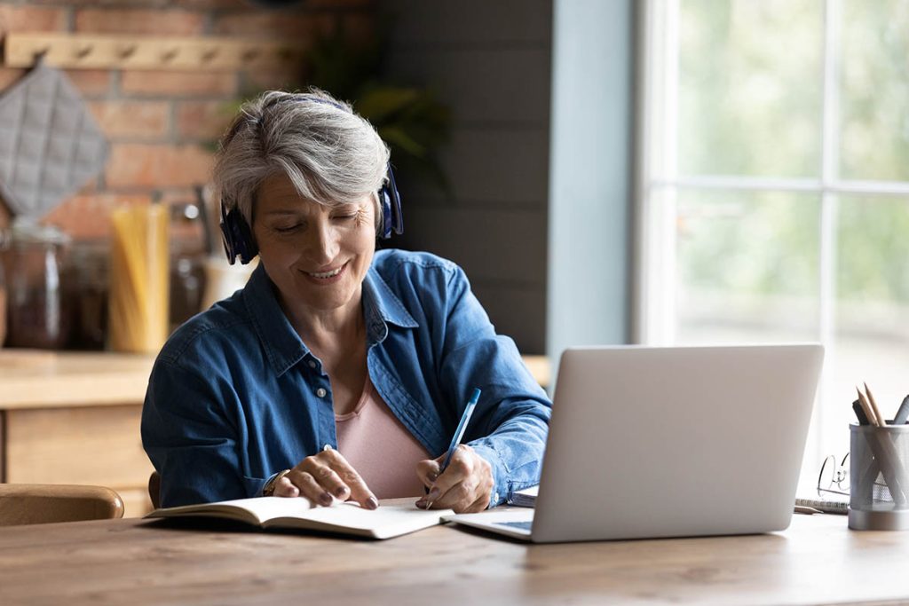 mature woman with headphones taking notes in front of her laptop