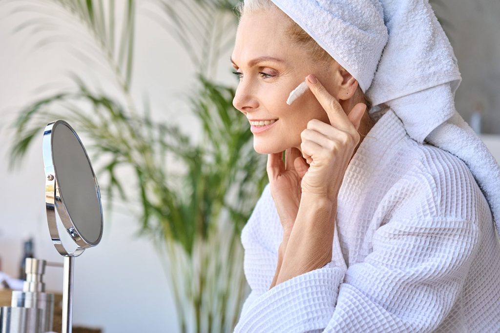 senior older adult 50 years old blonde woman wearing bathrobe and turban towel in bathroom applying moisturizing tightening face skin treatment, looking at mirror. Morning beauty routine.