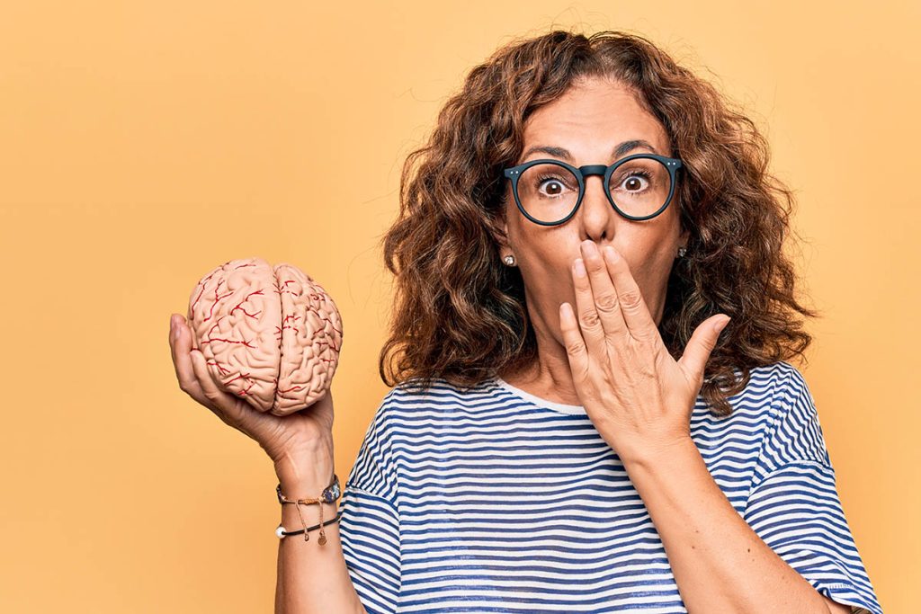 mature woman holding a brain with hand over her mouth in front of orange background