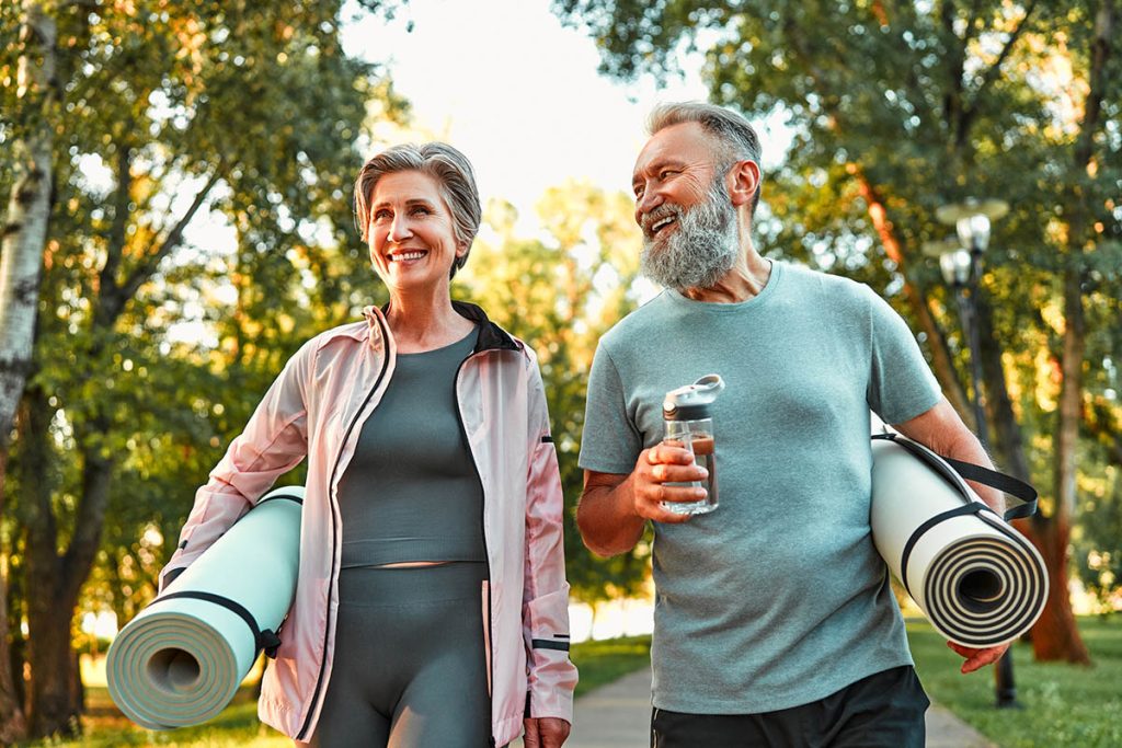 Active life of older people. Happy sports couple going for a workout outdoors, holding exercise mats and water.