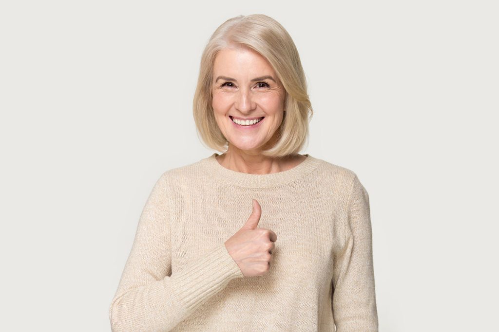 Smiling mature woman in sweater isolated on grey studio background show thumbs up recommend service, happy positive senior lady give sincere recommendation, advice good or sale offer