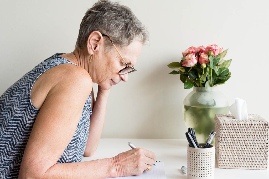 Profile view of older woman with short grey hair and glasses sitting at desk writing