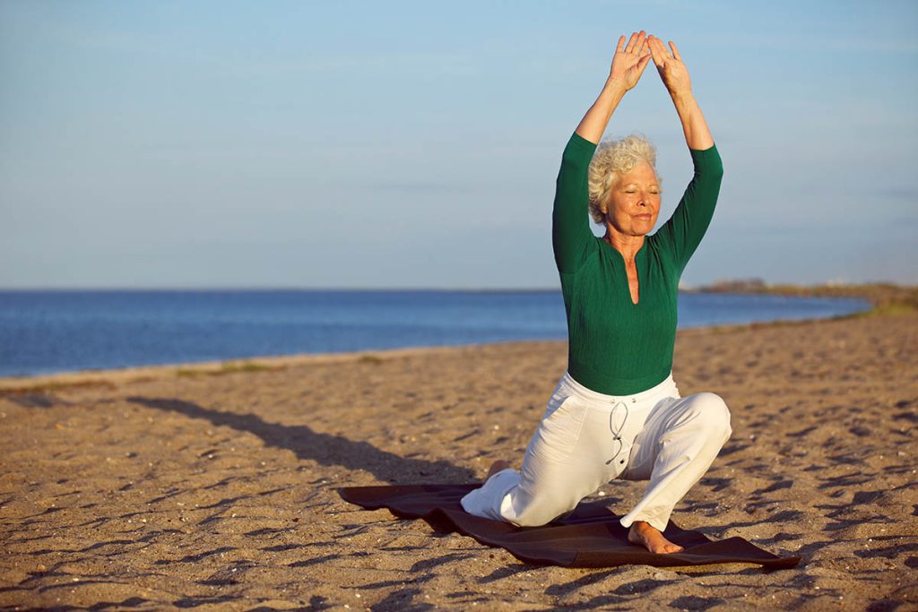 Senior woman practicing yoga poses on the sandy beach. Elder caucasian woman stretching legs and arms on the seashore. Healthy lifestyle and fitness concept