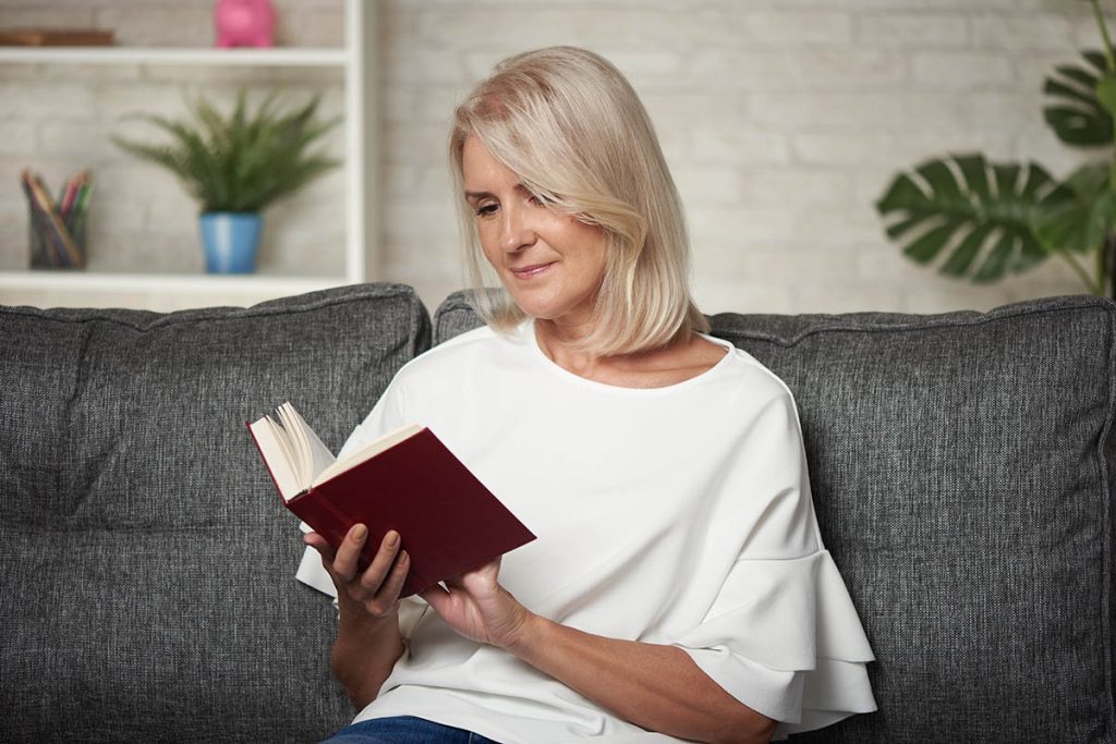 middle aged blonde woman is reading a book while sitting on a sofa at home