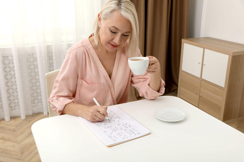 Middle aged woman with cup of drink solving sudoku puzzle at table indoors
