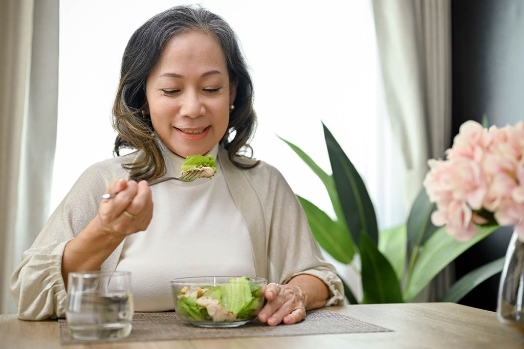 Beautiful and healthy Asian-aged woman eating healthy green salad with chicken breast in her dining room. Healthy and wellbeing lifestyle.