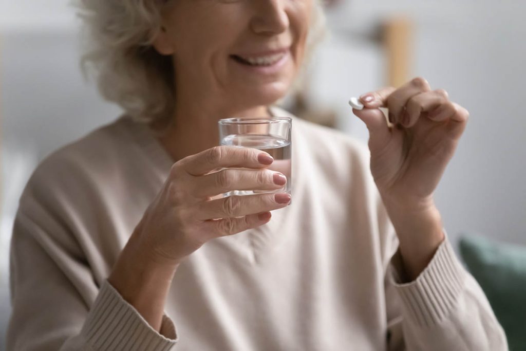 Smiling elderly female patient holding pill and glass of water. Senior mature woman taking daily dose of prescribed meds against depression, mental disorders, flu, insomnia, pain. Elderly treatment
