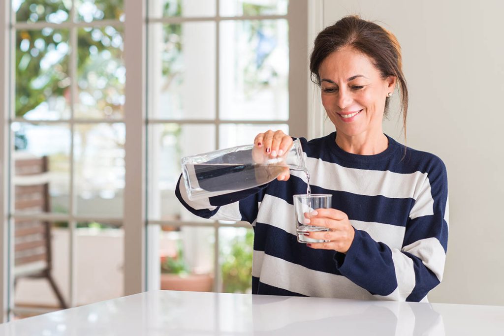 Middle aged woman drinking a glass of water with a happy face standing and smiling with a confident smile showing teeth