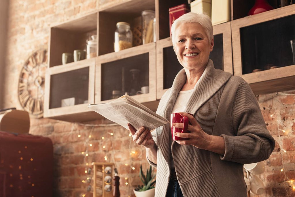 Aged Woman Reading Morning Newspaper And Drinking Coffee In Kitchen, Looking At Camera And Smiling