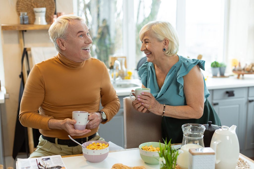 Happy couple having breakfast. Cheerful smiling glowing contended elderly grey-haired married couple wearing elegant clothing sharing a laugh while having breakfast at the kitchen