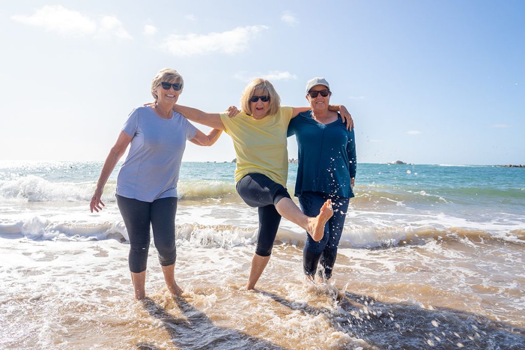 Lovely group of senior girl friends on their 60s walking and having fun splashing water on sea. Three mature healthy retired females Laughing and enjoying retirement and outdoors active lifestyle.