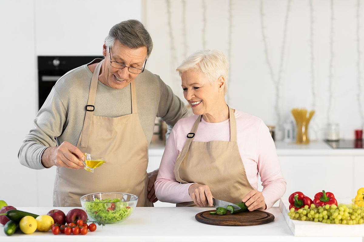 Happy Senior Couple Making Salad Together Cutting Vegetables And Dressing Dish In Kitchen. Elderly Spouses Cooking Dinner At Home. Modern Retirement Lifestyle And Healthy Nutrition Concept