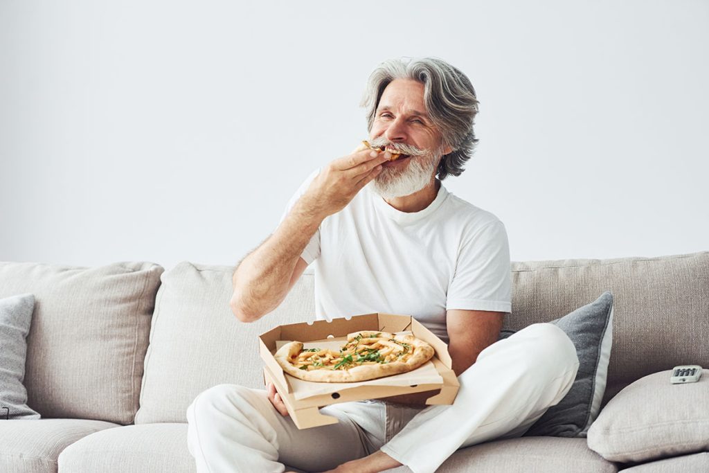 With delicious pizza. Watches TV show. Senior man with grey hair and beard indoors.